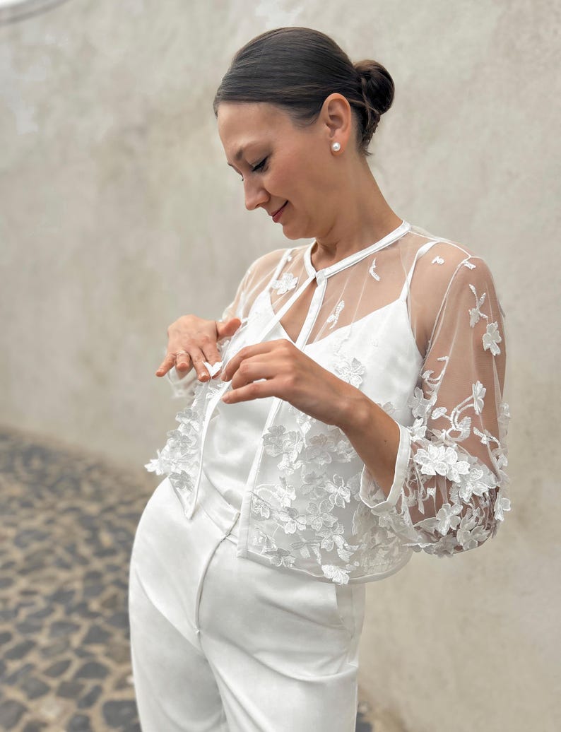 May include: A white lace bolero with a floral design. The bolero has a tie closure at the neck and sheer sleeves.