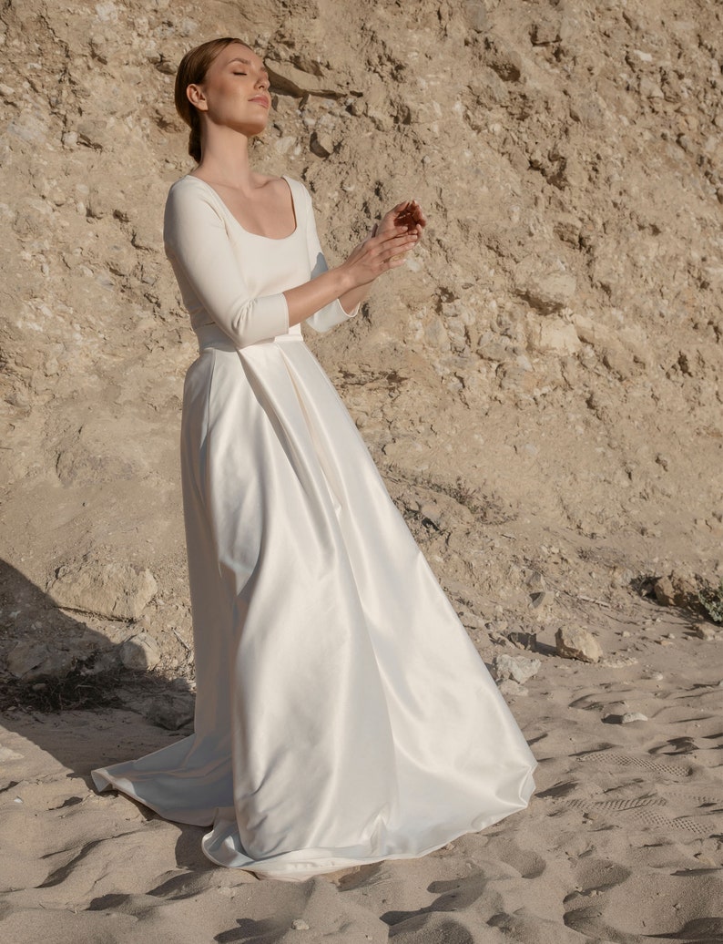 May include: A woman wearing a white, long-sleeved, scoop neck wedding dress with a satin skirt.