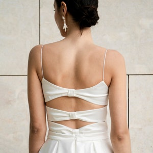 May include: A white satin wedding dress with a low back and a bow detail. The dress has a fitted bodice and a flowing skirt.