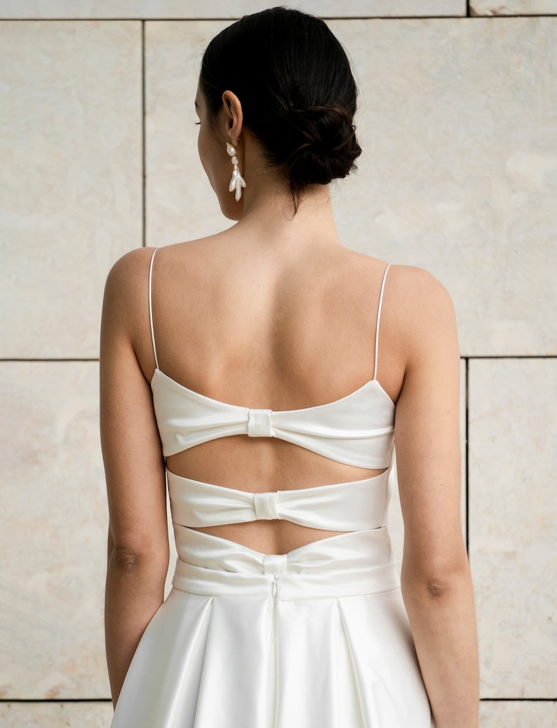 May include: A white satin wedding dress with a low back and a bow detail. The dress has a fitted bodice and a flowing skirt.