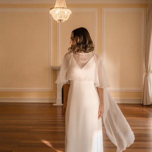 K&ouml;nnte beinhalten: Ein bodenlanges wei&szlig;es Kleid mit V-Ausschnitt und einem transparenten, flie&szlig;enden Cape-&Uuml;berwurf, der &uuml;ber die Schultern f&auml;llt und sich nach hinten erstreckt. Das Kleidungsst&uuml;ck besteht aus einem glatten, blickdichten Stoff mit schlichter Silhouette.