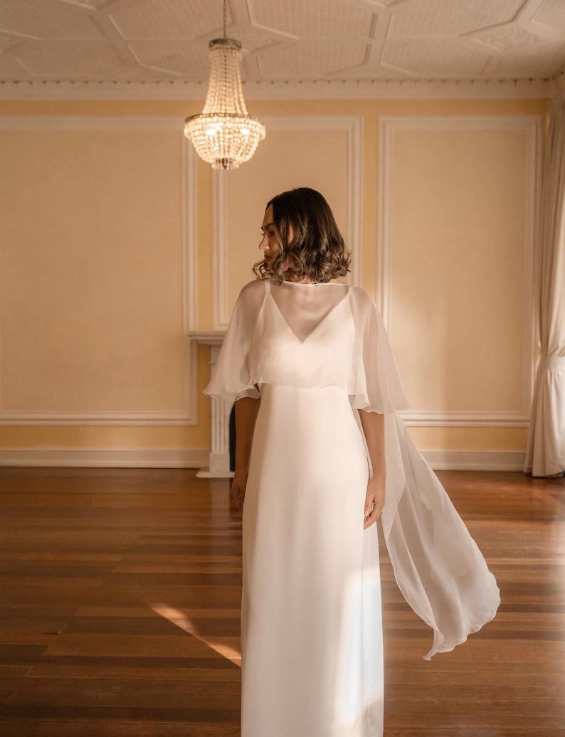 K&ouml;nnte beinhalten: Ein bodenlanges wei&szlig;es Kleid mit V-Ausschnitt und einem transparenten, flie&szlig;enden Cape-&Uuml;berwurf, der &uuml;ber die Schultern f&auml;llt und sich nach hinten erstreckt. Das Kleidungsst&uuml;ck besteht aus einem glatten, blickdichten Stoff mit schlichter Silhouette.