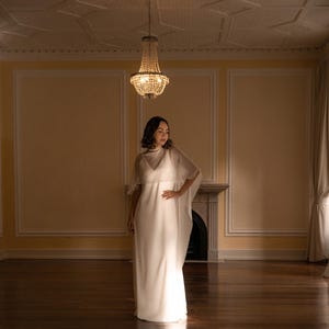 K&ouml;nnte beinhalten: Ein bodenlanges wei&szlig;es Kleid mit &auml;rmellosem Oberteil, V-Ausschnitt und einem transparenten, flie&szlig;enden Umhang, der &uuml;ber die Schultern und Arme f&auml;llt. Das Kleid besteht aus einem glatten, blickdichten Stoff und hat eine gerade Silhouette.