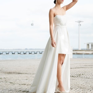 May include: A white high-low wedding dress with a fitted bodice and a flowing skirt. The dress has a simple design with a thin belt at the waist.