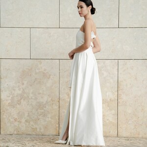 May include: A white high-low wedding dress with a spaghetti strap design and a slit in the skirt.