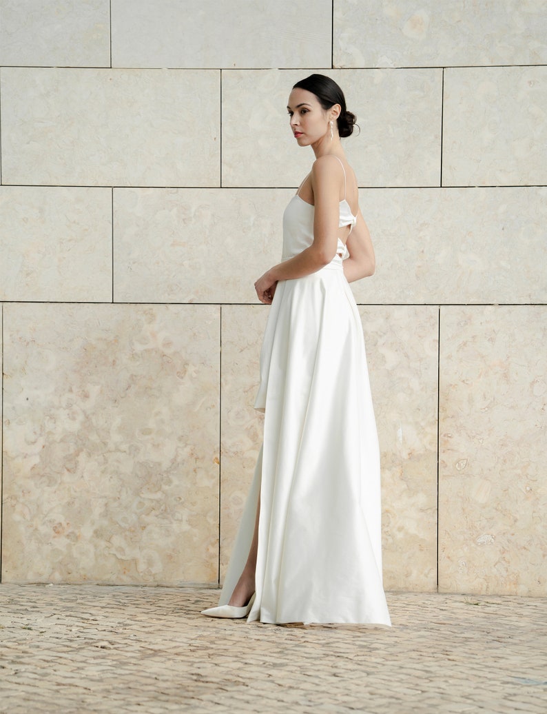 May include: A white high-low wedding dress with a spaghetti strap design and a slit in the skirt.