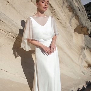 May include: A white wedding dress with a sheer, cape-like overlay. The dress has a V-neckline and a flowing skirt. The model is standing in front of a light-colored rock formation.