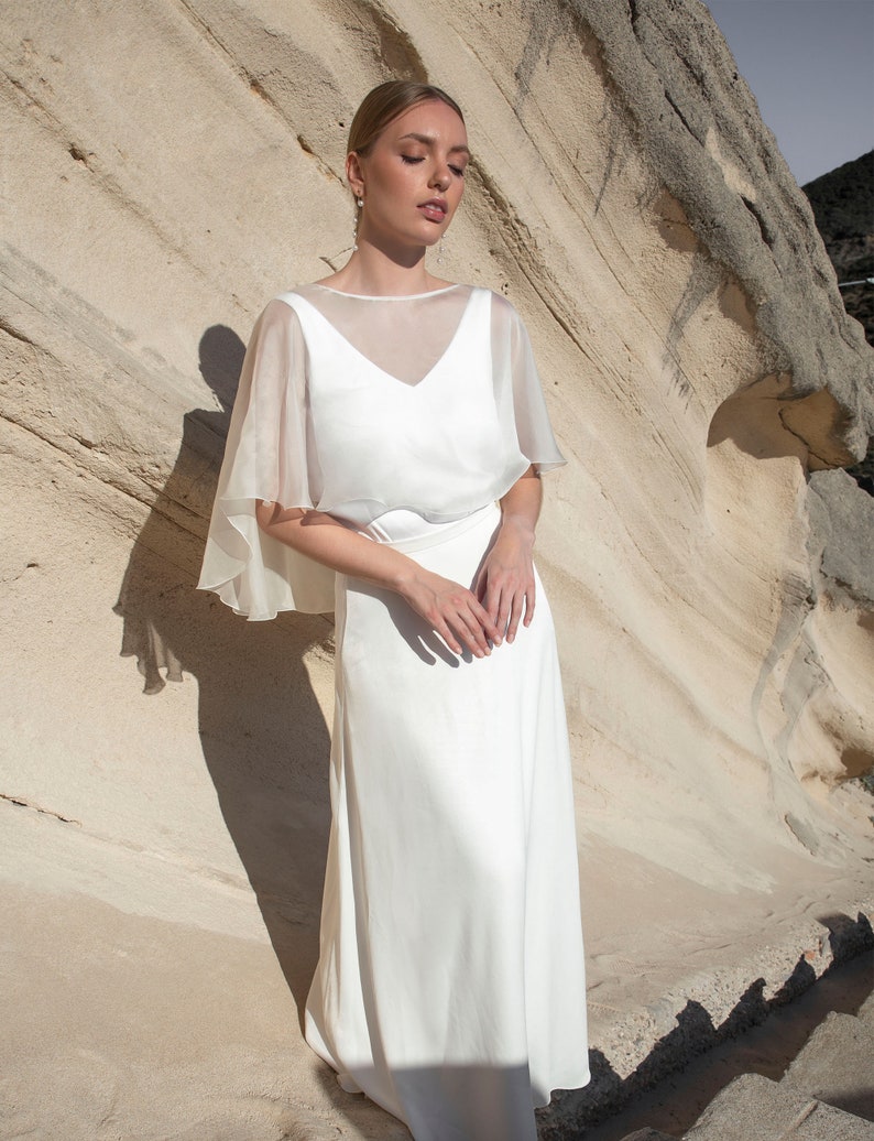May include: A white wedding dress with a sheer, cape-like overlay. The dress has a V-neckline and a flowing skirt. The model is standing in front of a light-colored rock formation.