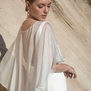 May include: A white, flowing cape with a layered design, worn over a white dress. The cape has a sheer, lightweight fabric and a rounded neckline. The model is wearing pearl earrings. The background is a light brown rock formation.