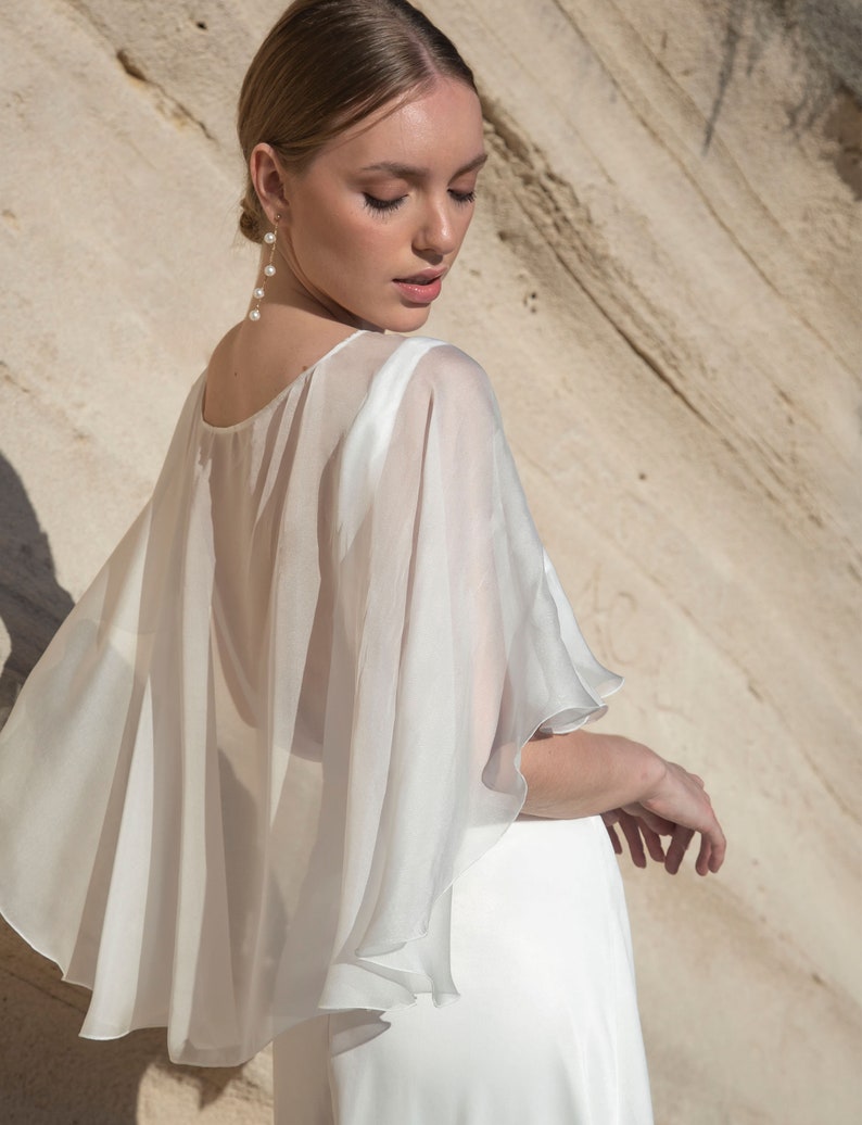 May include: A white, flowing cape with a layered design, worn over a white dress. The cape has a sheer, lightweight fabric and a rounded neckline. The model is wearing pearl earrings. The background is a light brown rock formation.