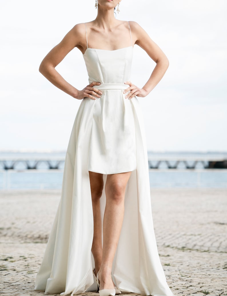 May include: A white sleeveless wedding dress with a short skirt and a long train. The dress has a simple design with a square neckline and a fitted bodice. The train is made of the same fabric as the skirt and flows gracefully behind the model.