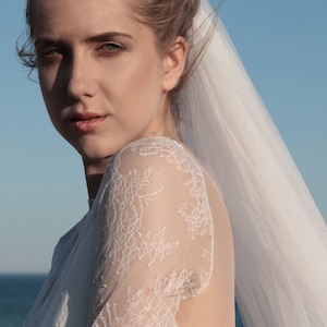 Puede incluir: Una novia con un vestido de novia de encaje blanco con velo. El vestido tiene la espalda baja y mangas largas. La novia est&aacute; mirando a la c&aacute;mara.