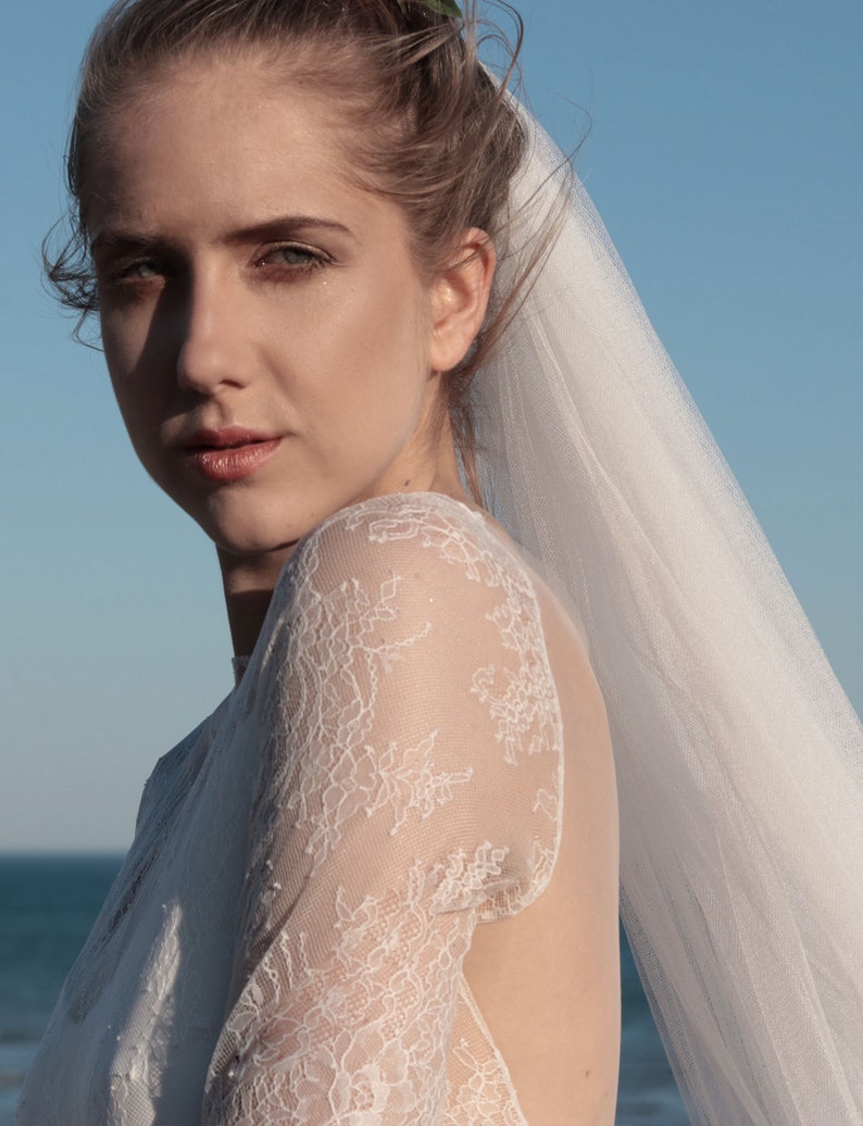 Puede incluir: Una novia con un vestido de novia de encaje blanco con velo. El vestido tiene la espalda baja y mangas largas. La novia est&aacute; mirando a la c&aacute;mara.