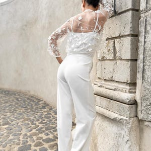 May include: A white jumpsuit with a sheer lace top featuring floral embellishments. The jumpsuit has a wide-leg silhouette and a fitted bodice.