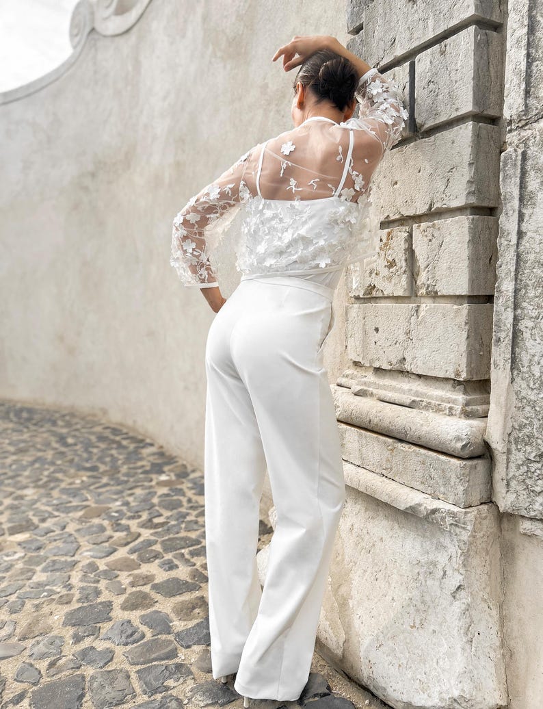 May include: A white jumpsuit with a sheer lace top featuring floral embellishments. The jumpsuit has a wide-leg silhouette and a fitted bodice.