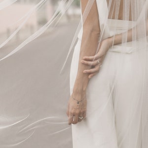 Puede incluir: Un velo de novia blanco de tejido delicado y transparente. El velo est&aacute; drapeado sobre la espalda de la novia, creando un look fluido y elegante.