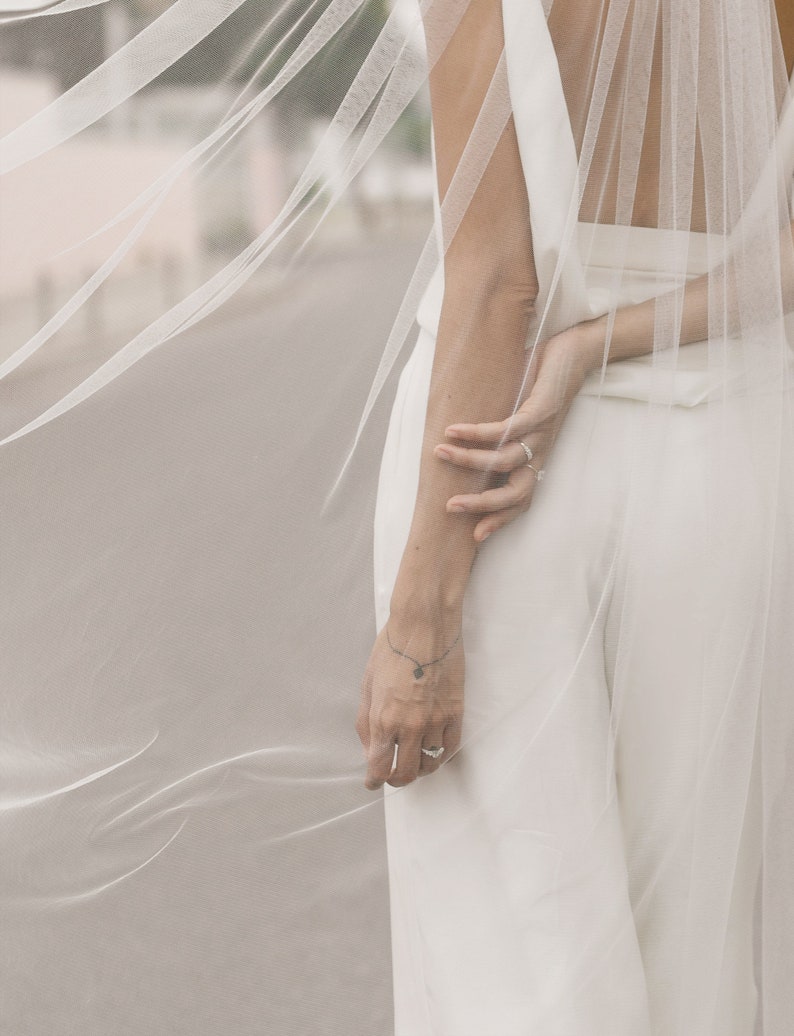 Puede incluir: Un velo de novia blanco de tejido delicado y transparente. El velo est&aacute; drapeado sobre la espalda de la novia, creando un look fluido y elegante.