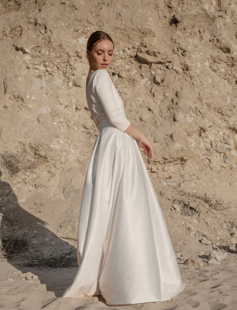 May include: A white satin wedding dress with a long skirt and a fitted bodice. The dress has a scoop neckline and three-quarter sleeves.