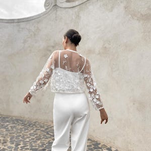 May include: A white sheer top with floral embroidery and long sleeves. The top is paired with white trousers.