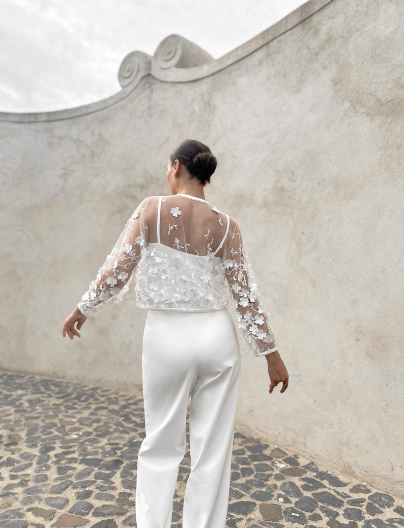 May include: A white sheer top with floral embroidery and long sleeves. The top is paired with white trousers.