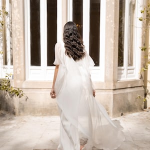 May include: A flowing white bridal gown with a cape detail. The dress is made of a lightweight fabric that drapes elegantly. The person is walking away from the camera, with long dark hair. The background features a large window.