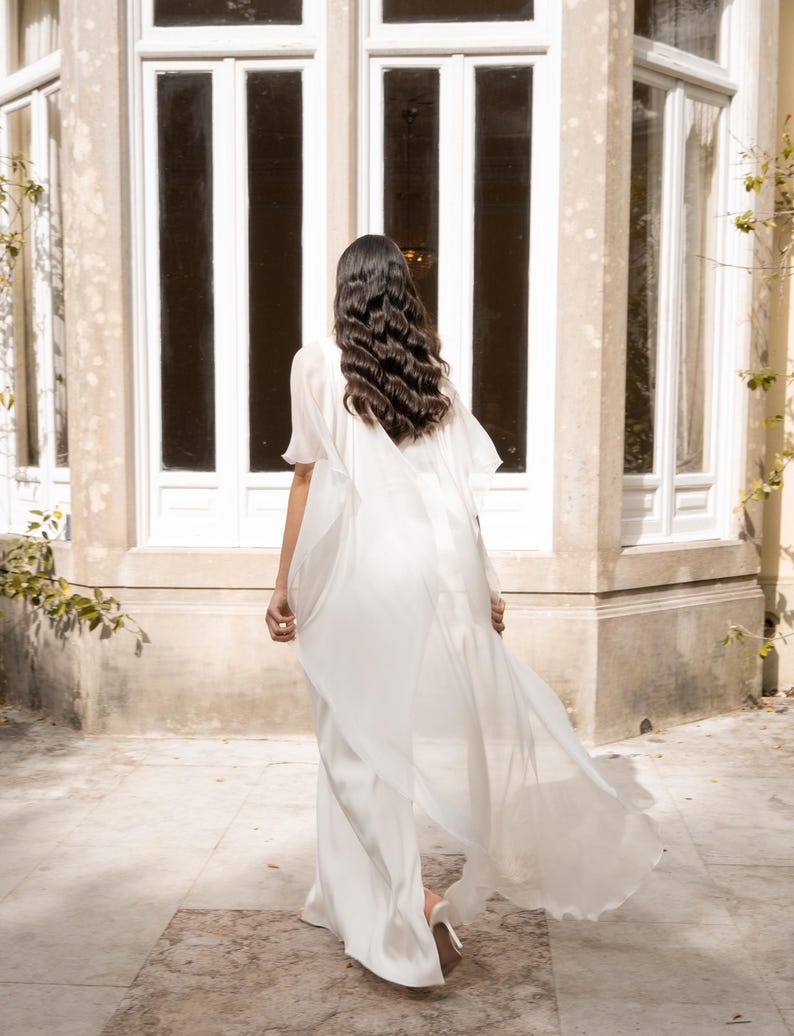 May include: A flowing white bridal gown with a cape detail. The dress is made of a lightweight fabric that drapes elegantly. The person is walking away from the camera, with long dark hair. The background features a large window.