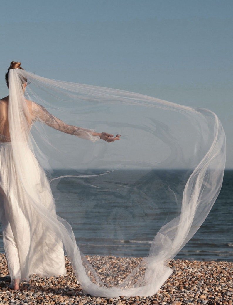 Puede incluir: Una novia con un vestido de novia blanco y un velo largo y fluido. El velo se mueve con el viento mientras ella est&aacute; de pie en una playa.