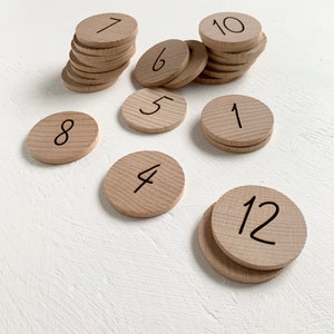 Könnte beinhalten: Holzscheiben mit den Zahlen 1, 4, 5, 6, 7, 8, 10, 12 und zwei 1er. Die Scheiben sind in Stapeln unterschiedlicher Höhe gestapelt.
