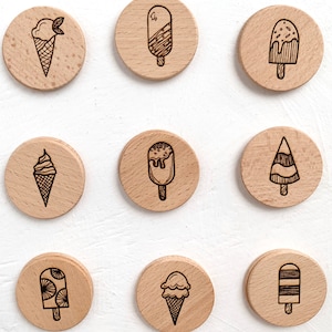 May include: Nine wooden round ornaments with different ice cream designs. The designs include a cone with a scoop of ice cream, a popsicle with sprinkles, a popsicle with a swirl design, a popsicle with a striped design, a popsicle with a geometric design, and a popsicle with a tree design.