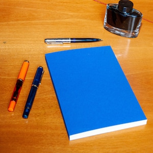 Könnte beinhalten: Ein leuchtend blaues Notizbuch in der Mitte, begleitet von drei Füllfederhaltern in Orange, Dunkelblau und Silber. Ein gläsernes Tintenfass mit schwarzem Deckel und ein silberner digitaler Musikplayer sind in der Nähe auf der Holzoberfläche platziert.