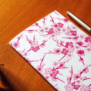 May include: A notebook with a white cover featuring a pink cherry blossom pattern, a silver and black pen, and a dark ink bottle rest on a wooden surface. The notebook's floral design is detailed and vibrant.