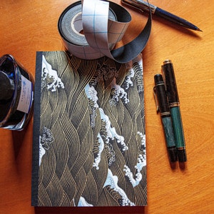May include: A black notebook with a gold and white wave pattern on the cover. Two green and black pens and a roll of black tape with a grid pattern are on a wooden surface.