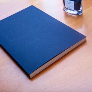 May include: A rectangular notebook with a solid dark blue cover and white pages. Two fountain pens with reddish-brown barrels and gold-colored accents are positioned to the left, and a glass ink bottle is visible near the top right corner.