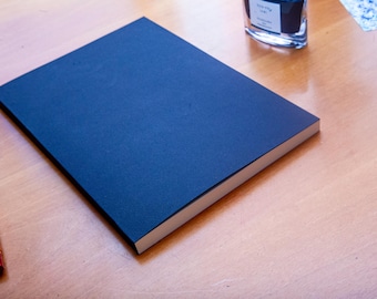 May include: A rectangular notebook with a solid dark blue cover and white pages. Two fountain pens with reddish-brown barrels and gold-colored accents are positioned to the left, and a glass ink bottle is visible near the top right corner.