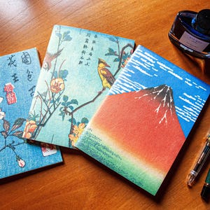 Può includere: Tre piccoli quaderni colorati con disegni di ispirazione giapponese. I quaderni presentano un design floreale blu, un design blu e verde con un uccello e fiori, e un design blu e rosso con una montagna.