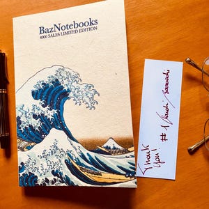 May include: A cream-colored notebook titled BazNotebooks 4000 SALES LIMITED EDITION featuring a blue wave illustration, accompanied by a blue fountain pen, a black striped fountain pen, a white note with handwritten text, and gold-rimmed glasses.