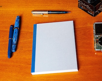 May include: A light gray notebook with a blue spine, two blue fountain pens, a silver and black ballpoint pen, a glass bottle of Edelstein ink, and a small silver digital music player with a black circular control pad and visible circuitry.