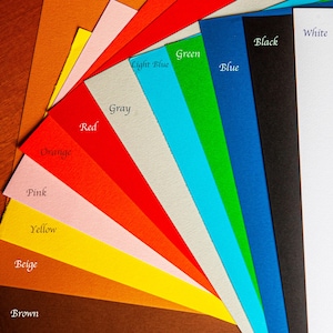 May include: A fan-shaped arrangement of rectangular paper sheets in various colors including brown, beige, yellow, pink, orange, red, gray, light blue, green, blue, black, and white. Each color is labeled with its corresponding name in black text.