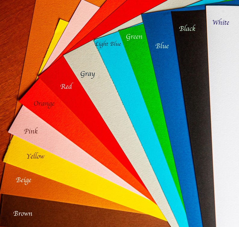 May include: A fan-shaped arrangement of rectangular paper sheets in various colors including brown, beige, yellow, pink, orange, red, gray, light blue, green, blue, black, and white. Each color is labeled with its corresponding name in black text.