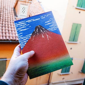 Puede incluir: Un lienzo rectangular que muestra una montaña estilizada con una cima oscura, una ladera en degradado rojo y naranja, y una base verde sobre un cielo azul con rayas de nubes horizontales blancas, todo sobre una superficie de lienzo texturizada.