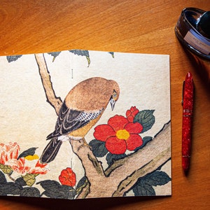 May include: An open notebook with a bird and floral illustration, a red pen, and a bottle of blue ink on a wooden surface. The notebook features a bird perched on a branch with red flowers.