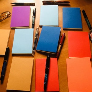 May include: A collection of nine rectangular notebooks in various solid colors including purple, light blue, dark blue, orange, and red, arranged with several fountain pens, a pair of eyeglasses, and a glass ink bottle with a black cap.