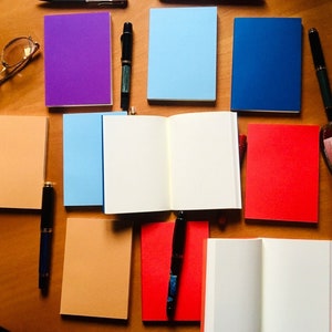 May include: A collection of rectangular notebooks in solid colors including purple, light blue, dark blue, peach, and red, arranged with several black fountain pens, a pair of gold-rimmed eyeglasses, and a glass ink bottle with a black cap.