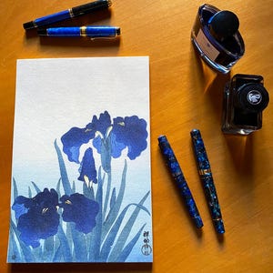Pode incluir: Uma natureza morta artística com canetas-tinteiro azuis, frascos de tinta e uma aguarela de flores de íris azuis. As canetas têm detalhes dourados. A pintura e as canetas estão dispostas sobre uma superfície de madeira.