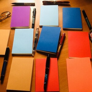 A top-down view of ten assorted colorful passport notebooks laid out on a wooden table along with various fountain pens, spectacles, and a glass inkwell.