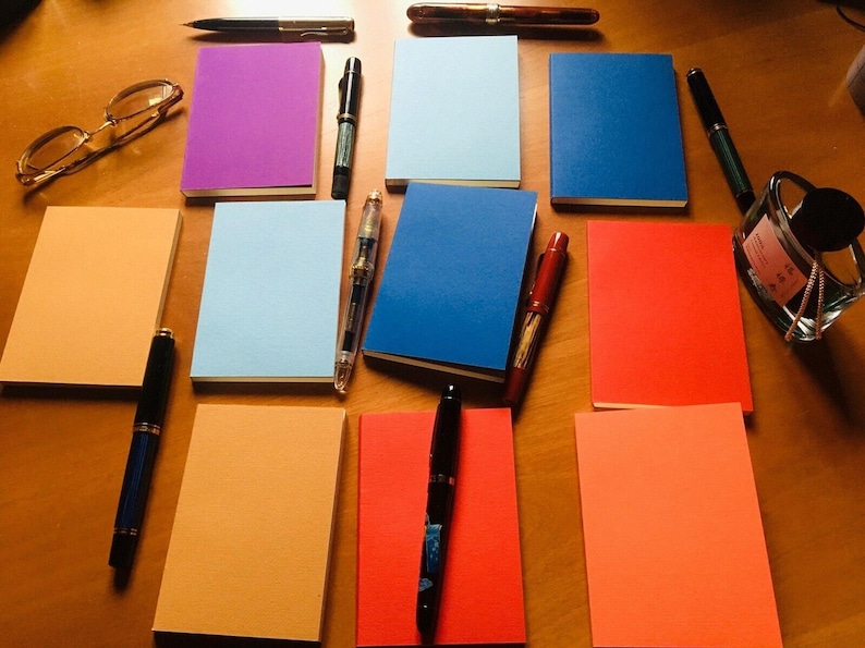 A top-down view of ten assorted colorful passport notebooks laid out on a wooden table along with various fountain pens, spectacles, and a glass inkwell.