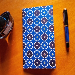 May include: A blue patterned notebook, a blue and black fountain pen, and a bottle of ink sit on a wooden surface. The notebook has a geometric design in white, blue, and red. The pen is blue with gold accents.