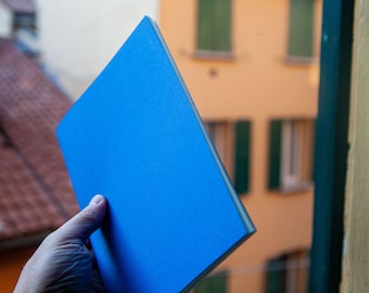 May include: A rectangular notebook with a solid, vibrant blue cover. The notebook is held at an angle, showcasing its clean, sharp edges and smooth, matte texture. The spine is thin and light-colored, contrasting with the bold blue front cover.