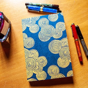 May include: A blue notebook with a gold spiral pattern. The notebook is on a wooden surface with three pens beside it. One pen is red, one is black, and one is blue.