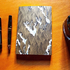 May include: A collection of writing supplies on a wooden surface. The items include three black fountain pens, a patterned notebook with a wave design in gold and white, and a bottle of ink. The pens have gold accents and the notebook is rectangular.
